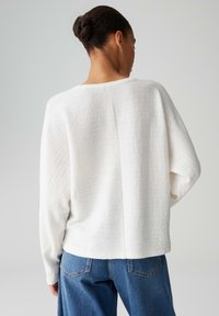 Donna con chignon intrecciato che indossa un maglione bianco a maniche lunghe con trama e jeans blu, vista di spalle su uno sfondo semplice.