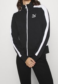 Sort zip-jakke med høj krave, hvide sidepaneler og Puma-logo. Har en tætsiddende design samt ribbede manchetter og kant.