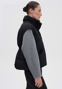 Schwarze Steppweste mit hohem Kragen und elastischer Taille, kombiniert mit einem hellgrauen Pullover. Glatte Textur, gestepptes Design, knielang.