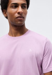 Giovane uomo che indossa una t-shirt viola chiaro con scollo rotondo e piccolo logo bianco sul petto, che guarda leggermente verso sinistra su uno sfondo semplice.