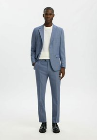 Costume bleu clair en tissu texturé, comprenant une veste unie boutonnage simple et un pantalon sur mesure, associé à un t-shirt blanc à col rond.