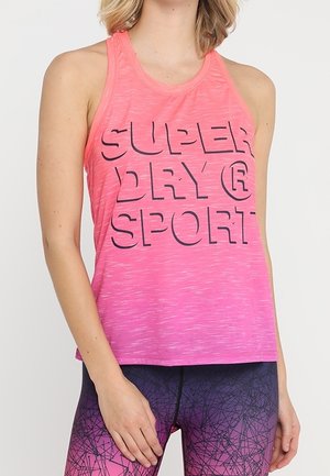 Kobieta ubrana w różowy sportowy top bez rękawów z przejściem kolorów i tekstem "SUPER DRY SPORT" oraz fioletowe legginsy w wzory na jednolitym tle.