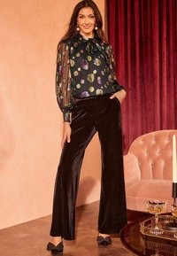 Pantaloni larghi neri in velluto abbinati a una blusa trasparente nera con pois colorati e scollo a cravatta. Accenti a fiocco neri sulle scarpe.