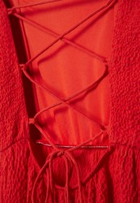 Punainen mekko, jossa on teksturoitu kangas, ristikkäin sidottu selkäosa ja nauhakiinnitys vyötäröllä, korostaen kerroksellista muotoilua.