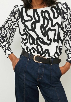 Frau trägt ein schwarz-weiß abstrakt gemustertes Langarmoberteil, in dunkelblaue hoch taillierte Jeans mit einem schwarzen Gürtel gesteckt.