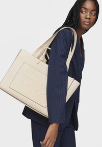 Sac fourre-tout en cuir beige avec logo embossé, forme structurée et larges anses avec un marquage ton sur ton. Porté par un modèle dans un costume à rayures.