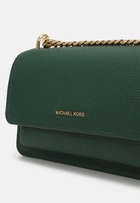 Zelená kožená kabelka s textura povrchem, zlatým řetízkem a vyraženým logem. Kompaktní obdélníkový tvar s klopou.