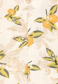 Let bluse med mønster af appelsinfrugter og grønne blade, med knaplukning og beige baggrund. Glat tekstur.