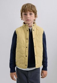 Quiltet gul vest med høj krave, trykknapper og en lomme, lagdelt over en mørkeblå striktrøje og en hvid skjorte.