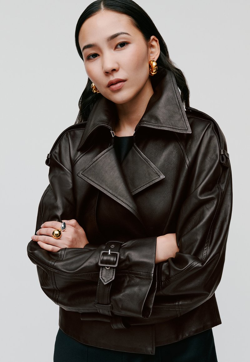 Braune Lederjacke mit breiten Revers, einer Schnalle in der Taille und strukturierten Ärmeln. Modell trägt goldene Ohrringe und einen Ring. Einfacher Hintergrund.