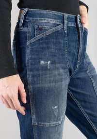 Jeans in denim di diverse sfumature di blu, con aree abrasate, diverse tasche e una vita standard con etichetta in pelle.