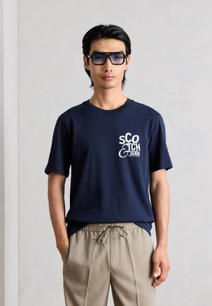Άνδρας που φοράει μπλε σκούρο μπλουζάκι Scotch & Soda, μπεζ παντελόνια με κορδόνια και μεγάλα σκουρόχρωμα γυαλιά ηλίου, στέκεται με τα χέρια στις τσέπες.