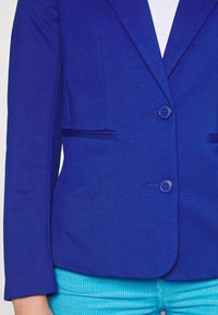 Blazer ajustado en tejido azul brillante con dos botones, dos bolsillos frontales con ribete sutil y corte entallado, combinado con pantalones turquesa.