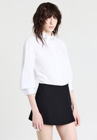 Giovane con capelli scuri bagnati che indossa una camicia bianca con bottoni e colletto decorato, accompagnata da una mini gonna nera, su uno sfondo bianco semplice.