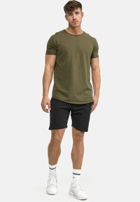 T-shirt olivastro a maniche corte con scollatura arrotondata abbinato a pantaloncini neri. Il modello indossa sneakers bianche e calzini alla coscia con strisce nere.