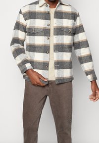 Veste à carreaux aux tons gris, crème et beige ; fabriquée en tissu doux. Comprend deux poches avant, une fermeture à boutons et un col classique.