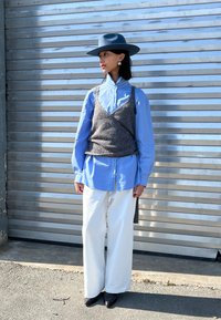 Grauer gestrickter Tanktop über einem hellblauen Hemd, kombiniert mit weiten weißen Hosen und schwarzen spitzen Absätzen. Mit einem Hut accessorisiert.