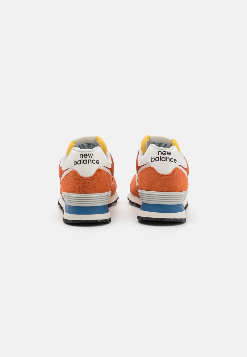 Baskets en daim orange avec des accents blancs, un onglet de talon jaune, une semelle intermédiaire bleue et une semelle extérieure noire. Logo "New Balance" affiché sur le talon.