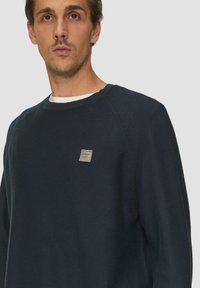 Navyblauer Pullover mit gerippter Textur, Rundhalsausschnitt und einem kleinen aufgenähten Etikett auf der Brust. Hergestellt aus weichem Stoff und mit Raglanärmeln.