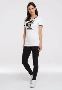 Weißes Baumwoll-T-Shirt mit schwarzem Besatz, das auf der Vorderseite eine cartoonhafte Maulwurf-Grafik zeigt. Kombiniert mit schwarzen Skinny-Jeans und weißen Sneakers.