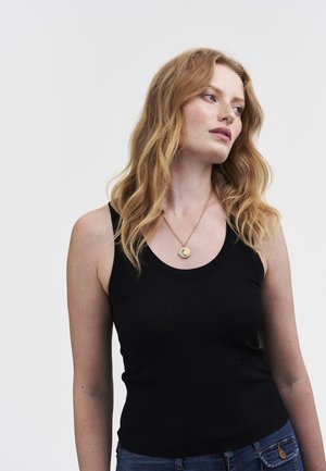 Jeune femme avec de longs cheveux bouclés rouges portant un haut sans manches noir, un jean bleu et un collier avec un pendentif en or, se tournant sur le côté.