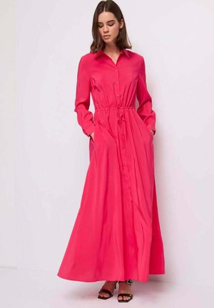 Donna che indossa un vestito maxi a maniche lunghe, rosa acceso, con bottoni e vita aderente, in piedi con le mani nelle tasche e tacchi neri aperti in punta.