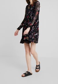 Schwarzes Blumenkleid mit langen transparenten Ärmeln, geraffter Taille und rüschiertem Saum. Kombiniert mit schwarzen Sandalen mit zwei Schnallen.