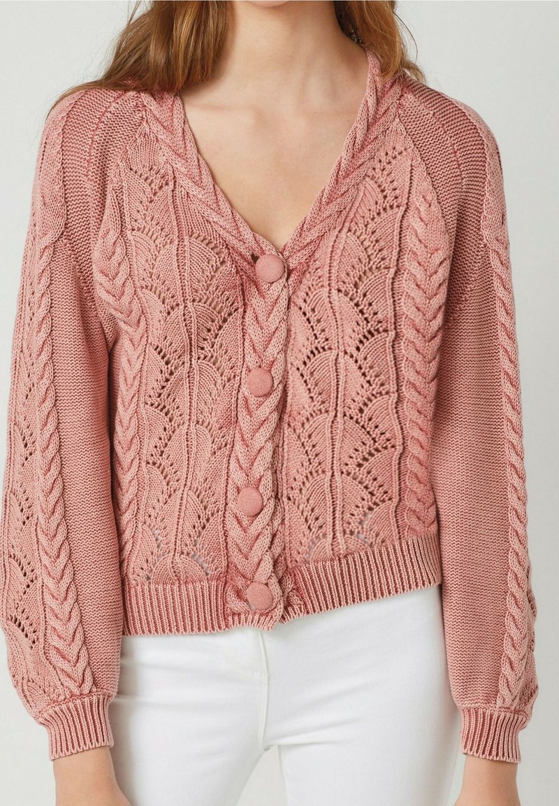 Donna che indossa un cardigan lavorato a maglia rosa, con motivi a treccia e pizzo, abbinato a pantaloni bianchi, su uno sfondo neutro.