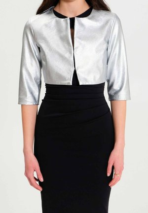 Kobieta ubranа w srebrną krótką kurtkę z rękawami do łokcia na dopasowanej czarnej sukience, pozująca na jednolitym jasnym tle.