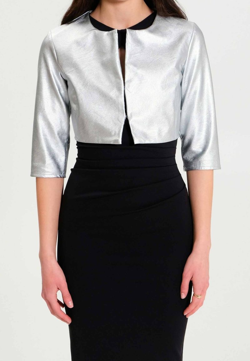 Femme portant une veste argentée courte à manches trois-quarts sur une robe noire ajustée, posant devant un fond uni clair.