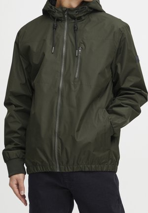 Mand iført olivengrøn hættetrøje med forreste lynlås og lynlåslomme på brystet, hænderne i sidelommerne, stående mod en hvid baggrund.