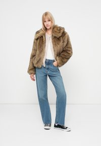 Giacca in pelliccia sintetica nelle tonalità marroni con una texture morbida, dotata di un ampio colletto. Abbinata a jeans azzurri chiari e sneakers nere.