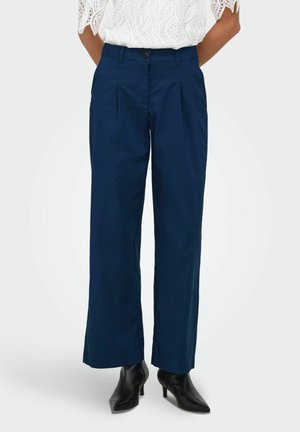 Vrouw die marineblauwe wijde broek met plooien draagt, gecombineerd met zwarte spitse haklaarzen en een witte kanten blouse.