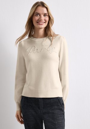 Femme souriante aux longs cheveux blonds porte un pull crème avec le texte "Paris" et un pantalon texturé foncé sur un fond clair uni.