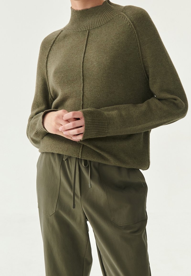 Personne portant un pull en maille côtelée vert olive à col haut et un pantalon assorti avec cordon, les mains doucement croisées à la taille.