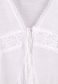 Blusa leggera bianca con pizzo decorato da motivi circolari, chiusura anteriore con nodo e tessuto testurizzato. Progettata per un fit rilassato.