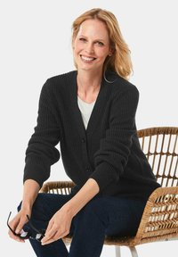 Schwarze gerippte Strickjacke mit V-Ausschnitt, Knopfschluss und umgeschlagenen Ärmel, getragen über einem weißen Hemd, kombiniert mit dunklen Jeans.