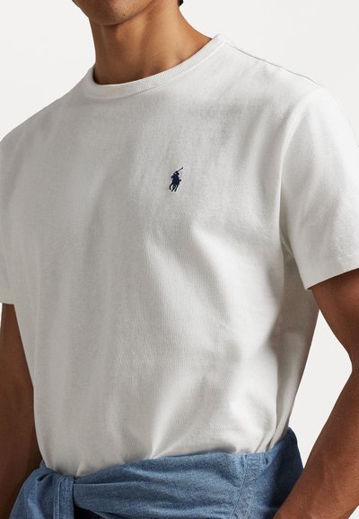 Λευκή βαμβακερή μπλούζα με στρογγυλή λαιμόκοψη και μικρό ναυτικό λογότυπο στο στήθος, συνδυασμένη με έναν ανοιχτό μπλε τζιν περιτύλιγμα δεμένο στη μέση.