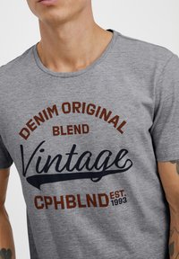 Šedé tričko s krátkým rukávem vyrobené z měkké tkaniny má grafický potisk s textem "DENIM ORIGINAL BLEND VINTAGE" v hnědé a námořnické barvě.