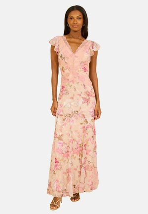 Kvinde iført en lang lyserød blomsterkjole med flagrende kapærmer og V-hals, kombineret med beige strop-sandaler, stående mod en hvid baggrund.