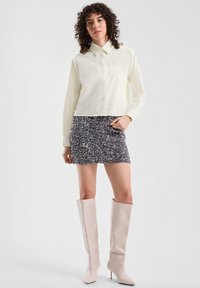 Przycięta jasnobeżowa koszula z guzikami w zestawieniu z błyszcząca mini spódnicą w kolorze srebrnym i szarym. Modelka nosi beżowe kozaki na wysokim obcasie do kolan.