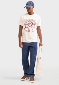 T-shirt creme com estampa vermelha de uma figura barbada, combinada com jeans azuis e ténis castanhos. Saco de lona na mão.