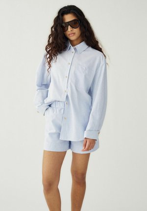 Kobieta z długimi kręconymi włosami, ubrana w oversize'ową jasnoniebieską koszulę w paski z guzikami, pasujące szorty i duże ciemne okulary przeciwsłoneczne, stoi z ręką w kieszeni.