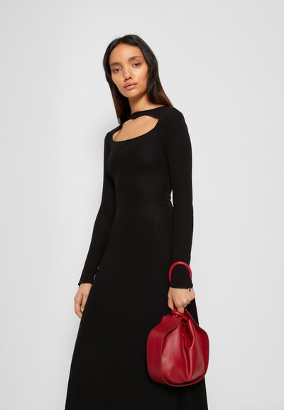 Vestido preto de manga longa com um decote quadrado. A modelo segura uma pequena bolsa de pele vermelha com uma alça curva.