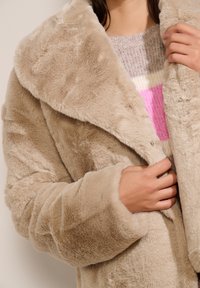 Tan nepbontenjas met een brede kraag, textuurstof en een knoopsluiting aan de voorkant. Details zijn onder andere zachte mouwen en een comfortabele binnenvoering.