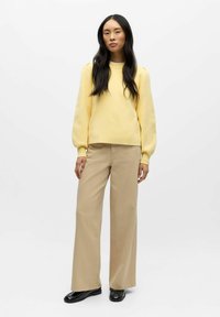 Žlutá mikina s nafouknutými rukávy, doplněná béžovými širokými kalhotami a černými botami. Hladká textura s volným střihem.