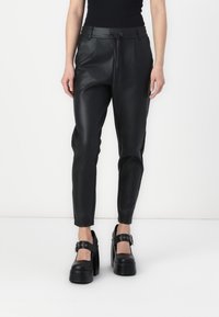 Vrouw in zwarte hoge faux leer broek met een trekkoord en zwarte platform Mary Jane schoenen tegen een witte achtergrond.