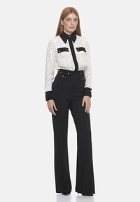 Blusa bianca con accenti neri, motivo a diamante e due tasche sul petto abbinata a pantaloni neri a vita alta con bottoni dorati e gambe svasate.