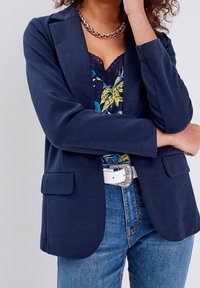 Veste bleu marine en matière douce avec une coupe ajustée. Dotée de deux poches avant et de revers crantés. Portée sur un haut à imprimé floral.