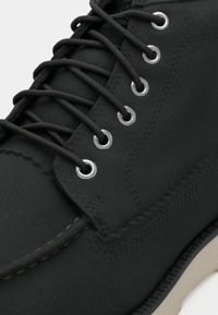 Black leather shoe with a smooth texture, featuring black laces, silver eyelets, and beige rubber sole. Stitching detail is visible along edges.